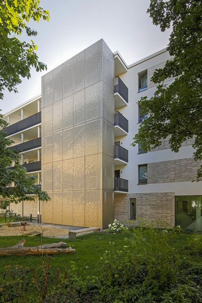 Perspektivische Außenansicht des Wohnkomplexes am Obentrautpark in Berlin. Im Vordergrund steht der markante, mit gold-bronzefarbenem Lochblech verkleidete Erschließungsturm. Die metallische Textur fängt das Sonnenlicht ein und bildet einen hochwertigen Akzent zur schlichten, weißen Putzfassade und den dunklen Balkongeländern des angrenzenden Hauptgebäudes.